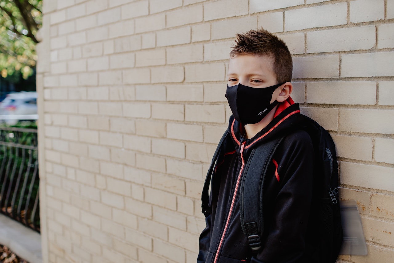young male student leaning against brick wall wearing a face mask
